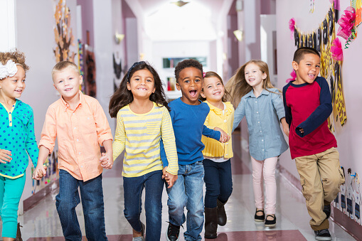 Seven elementary students hold hands and run joyfully through a school hallway.